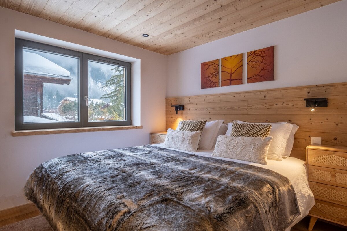 A warm bedroom features a large bed with a faux fur blanket, wood accents, and a window framing a serene snowy mountain view.