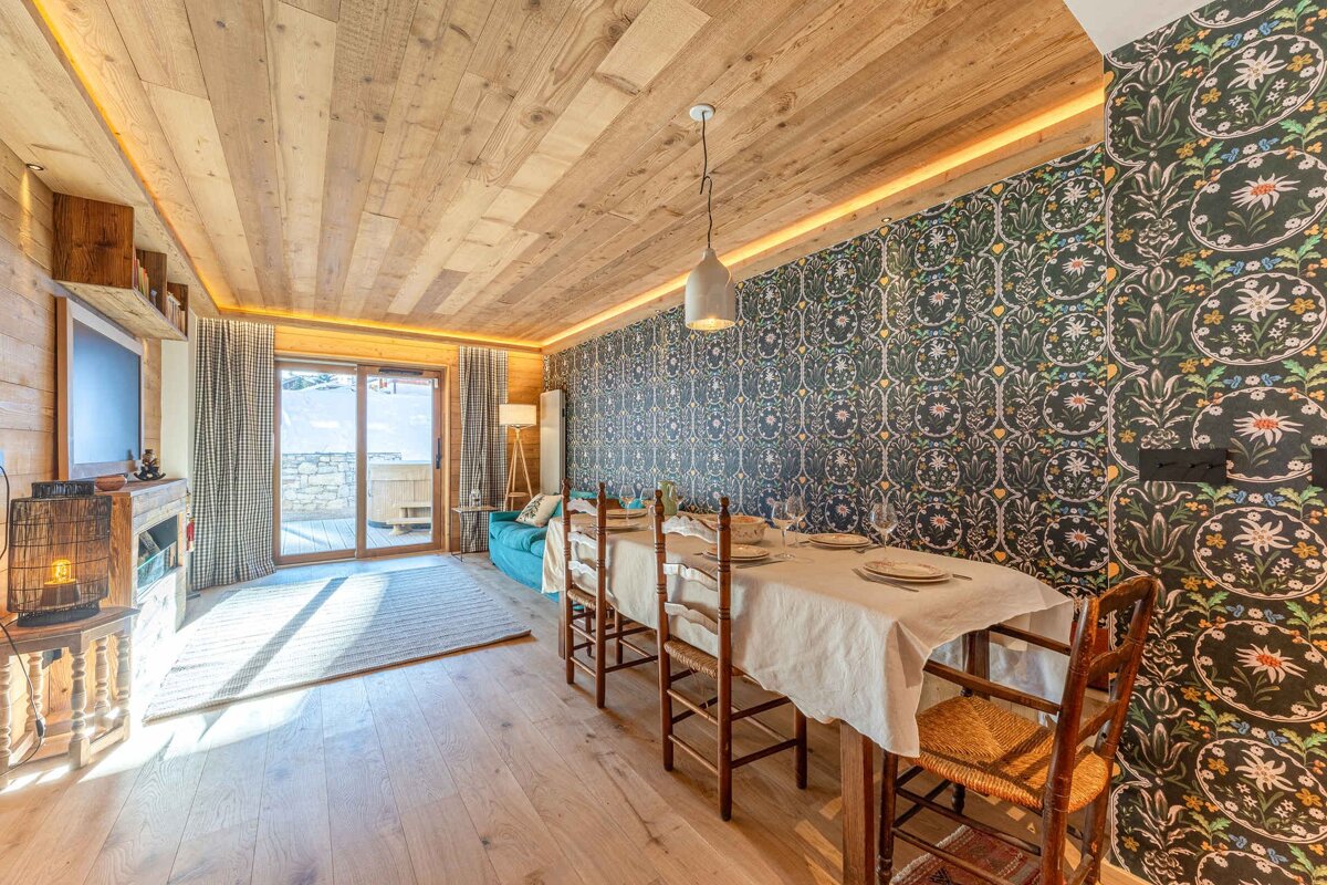 A warm, rustic dining and living space featuring wood paneling, dark floral wallpaper, a dining table, and a sunlit balcony.