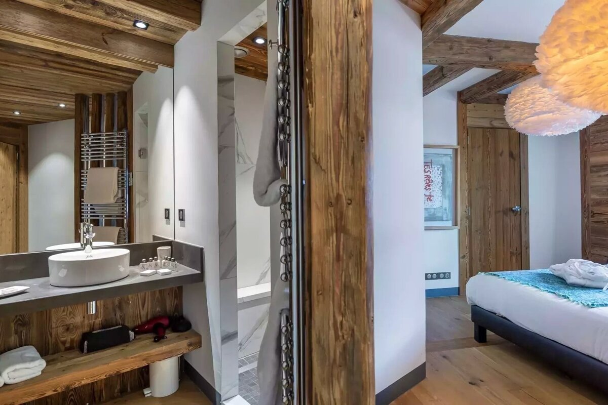 Cozy rustic-modern suite featuring an open bathroom with a round sink and shower, connected to a bedroom with a comfortable bed and wooden ceiling beams.