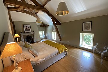 Warm attic bedroom with exposed wooden beams, a large bed with a yellow throw, a fireplace, small windows, and light wooden floors.