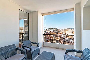 Sunny balcony with dark patio furniture and a panoramic view of a city with red-tiled roofs against a clear blue sky.