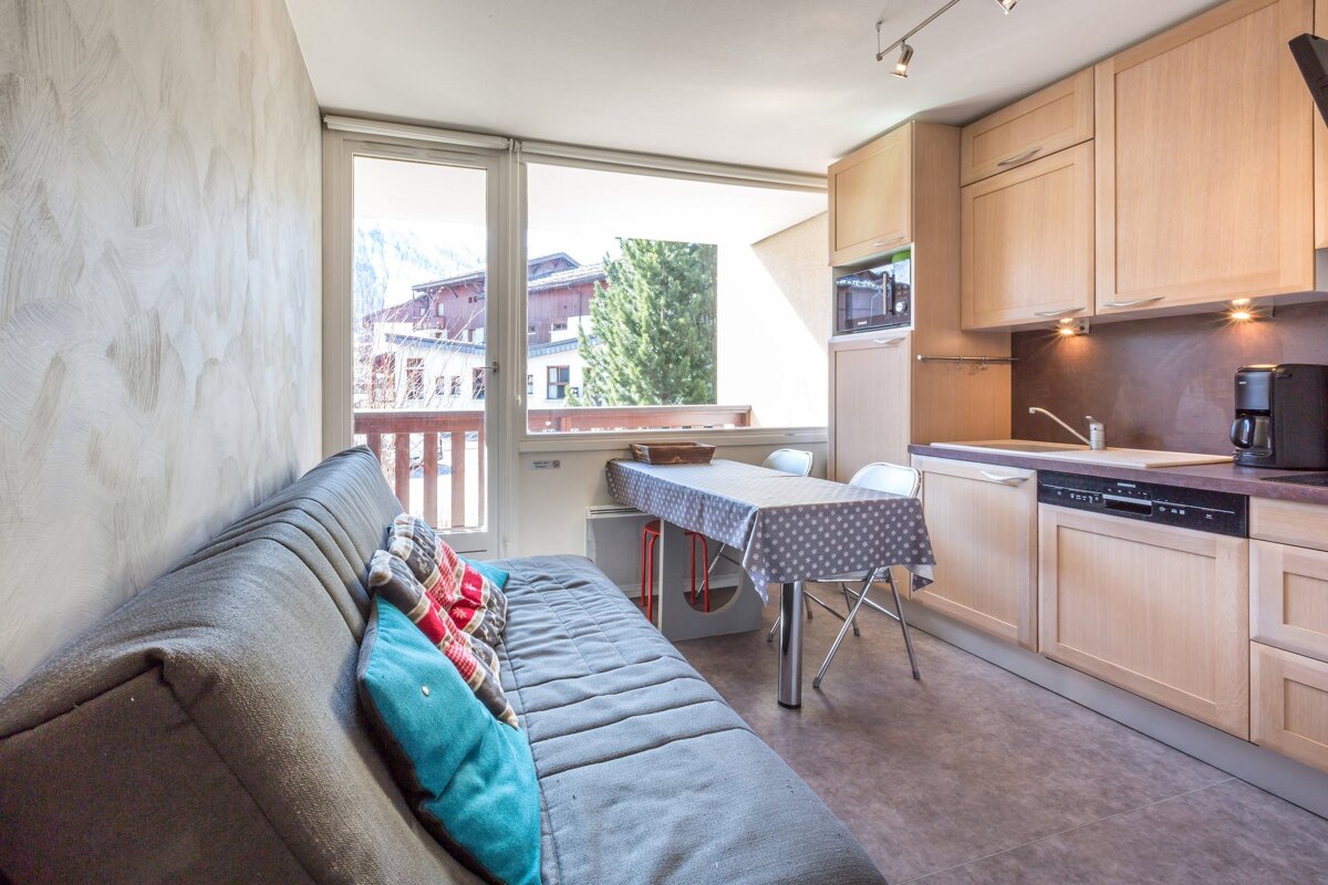 Cozy studio with a comfortable gray sofa, dining table, and a well-equipped kitchenette. Large windows offer a view of the surrounding mountain town.