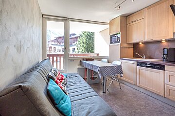 Cozy studio with a comfortable gray sofa, dining table, and a well-equipped kitchenette. Large windows offer a view of the surrounding mountain town.