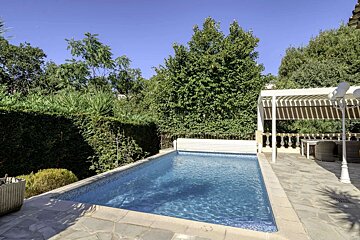 A rectangular swimming pool with blue water surrounded by stone paving and lush green trees. A white pergola with outdoor seating is visible on the right.