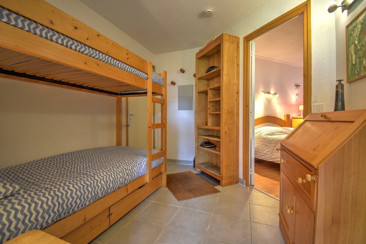 A room with wooden bunk beds, a wooden bookshelf, and a tiled floor, with a glimpse into an adjacent bedroom through an open doorway.