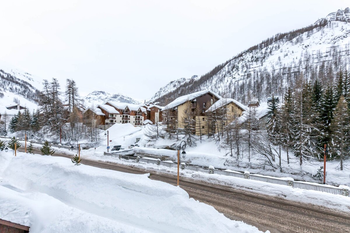 Snow-covered alpine chalets nestled in a winter mountain landscape with pine trees and a cleared road.