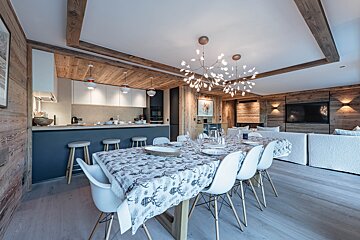 Modern chalet interior featuring a dining area with unique branch chandeliers, wood paneling, and a kitchen in the background.