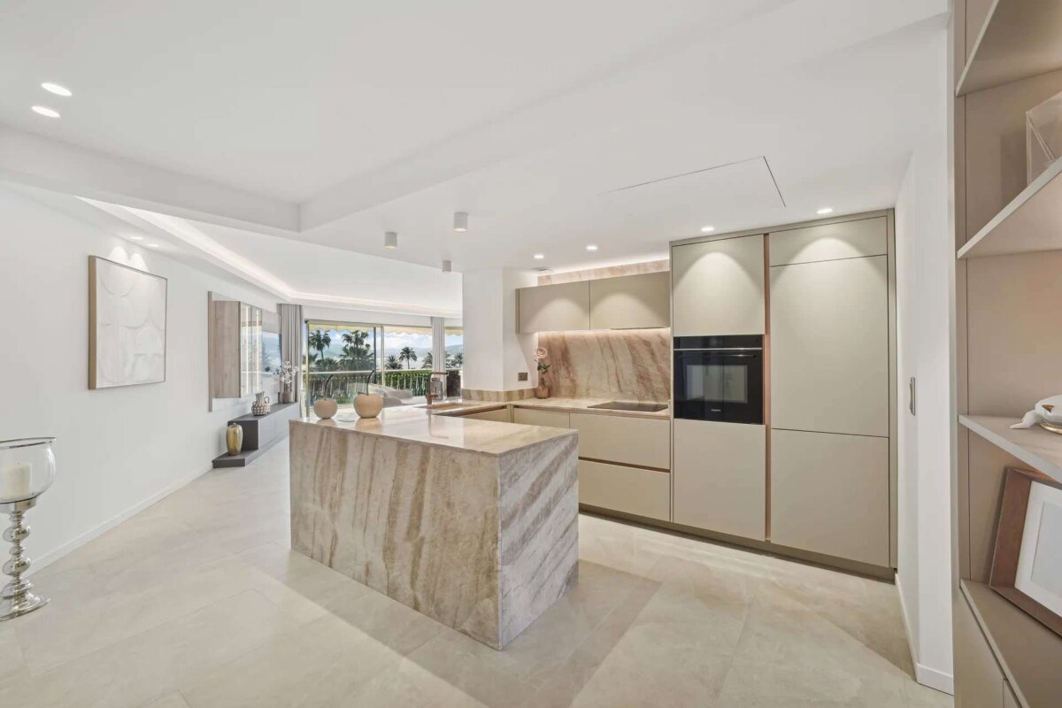 Bright, open-concept modern kitchen with a striking marble island and light cabinetry. It offers an expansive view of palm trees and outdoors.