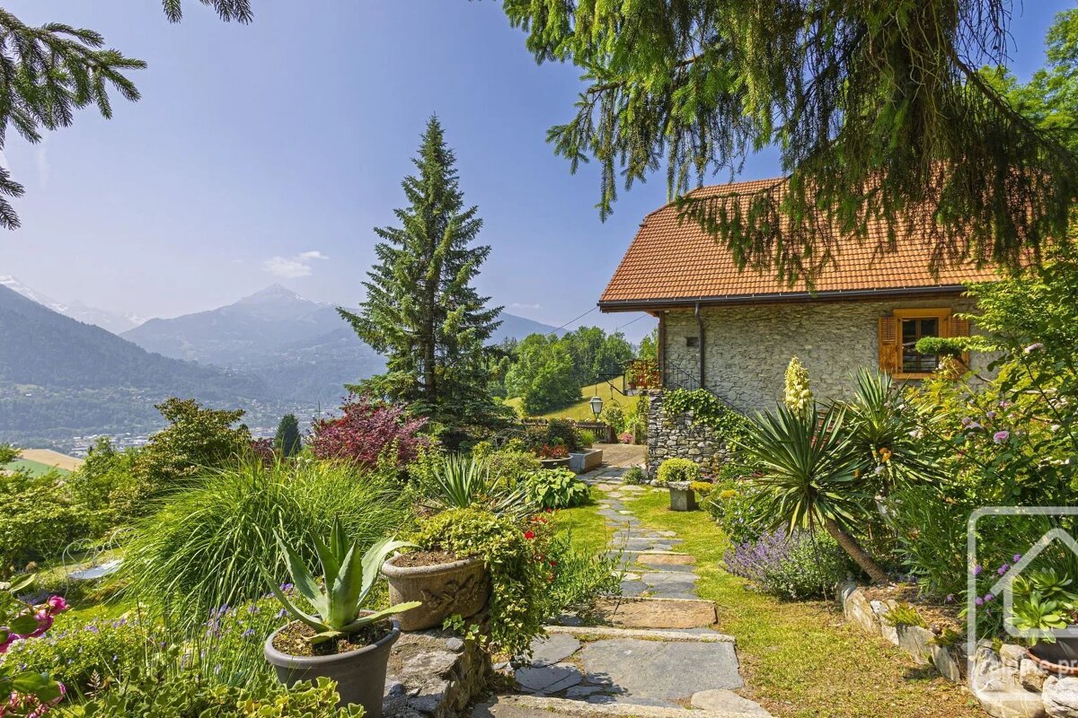 Cozy stone cottage with a lush, vibrant garden and stone path overlooking a breathtaking mountain valley under a clear blue sky.