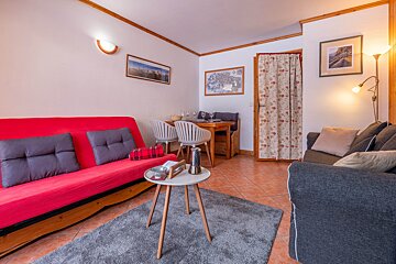 A cozy living/dining room featuring a red futon, gray sofa, wooden dining set, tiled floor, and warm lighting, creating an inviting space.