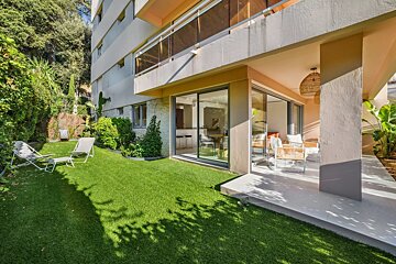 Ground-floor apartment with a lush artificial turf garden, lounge chairs, and a covered patio with modern seating and large glass doors.
