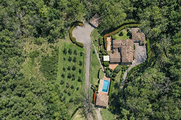 Aerial view of a secluded villa with red tile roofs, swimming pool, driveway, and orchard, nestled within a dense green forest.