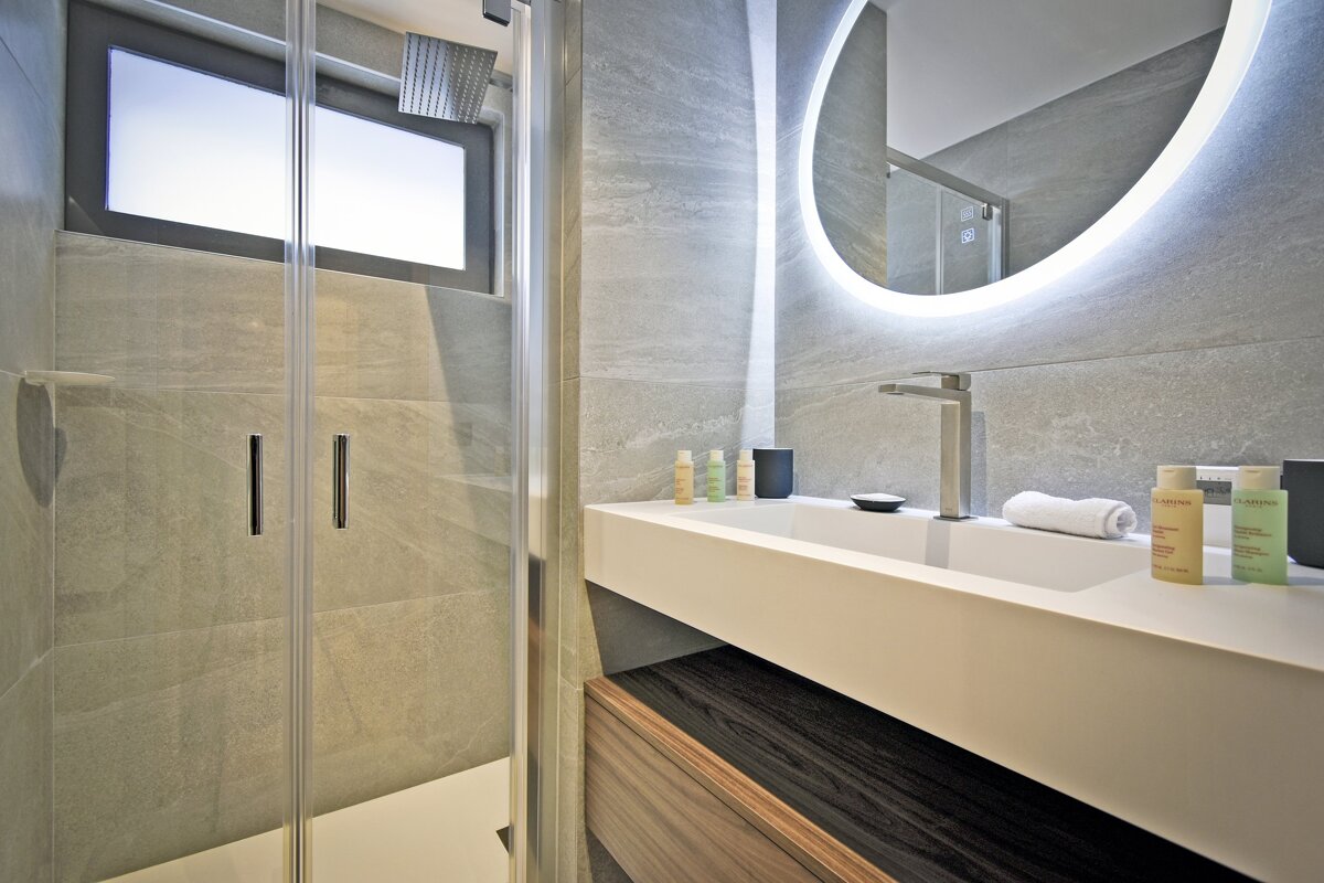 A modern bathroom features a glass shower, sleek white vanity with a rectangular sink, an illuminated round mirror, and grey tiled walls.