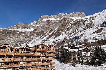 A scenic winter mountain resort with multi-story wooden chalets and traditional homes, set against towering, snow-covered peaks under a bright blue sky.