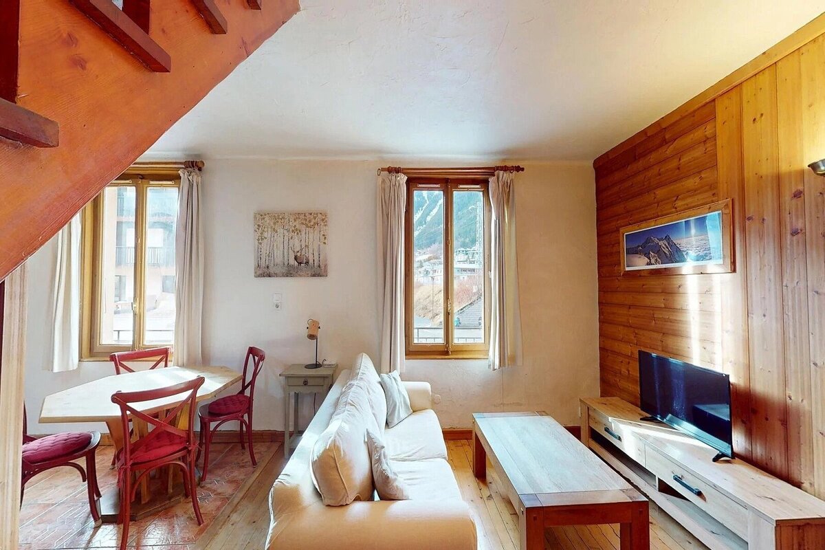 Cozy living/dining room with a white sofa, wooden table, and red chairs. Windows offer mountain views, complemented by wood paneling and accents.