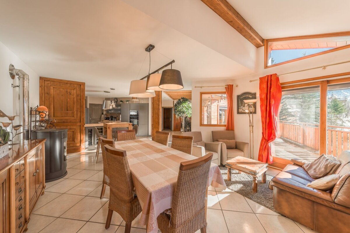 Bright open-plan chalet interior featuring a dining table with wicker chairs, a leather sofa, and glass doors to a balcony.