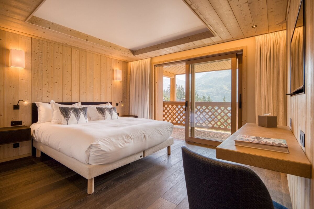 Cozy wood-paneled bedroom featuring a white bed, a desk, and sliding glass doors opening to a balcony with mountain views.