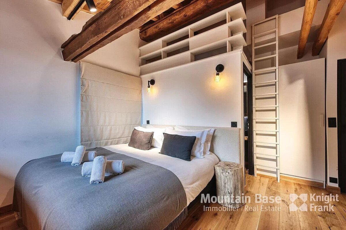 A cozy bedroom with a large bed, exposed wooden beams, built-in shelving, and a ladder, blending rustic and modern design.