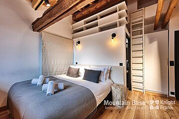 A cozy bedroom with a large bed, exposed wooden beams, built-in shelving, and a ladder, blending rustic and modern design.