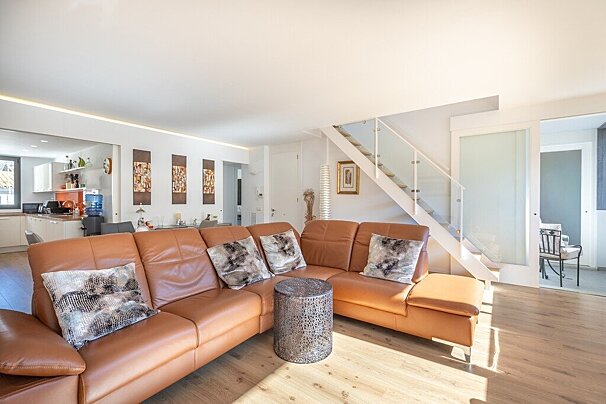 A bright, modern open-plan living room featuring a large brown leather sectional sofa, light wood floors, and a white staircase. A kitchen is visible.