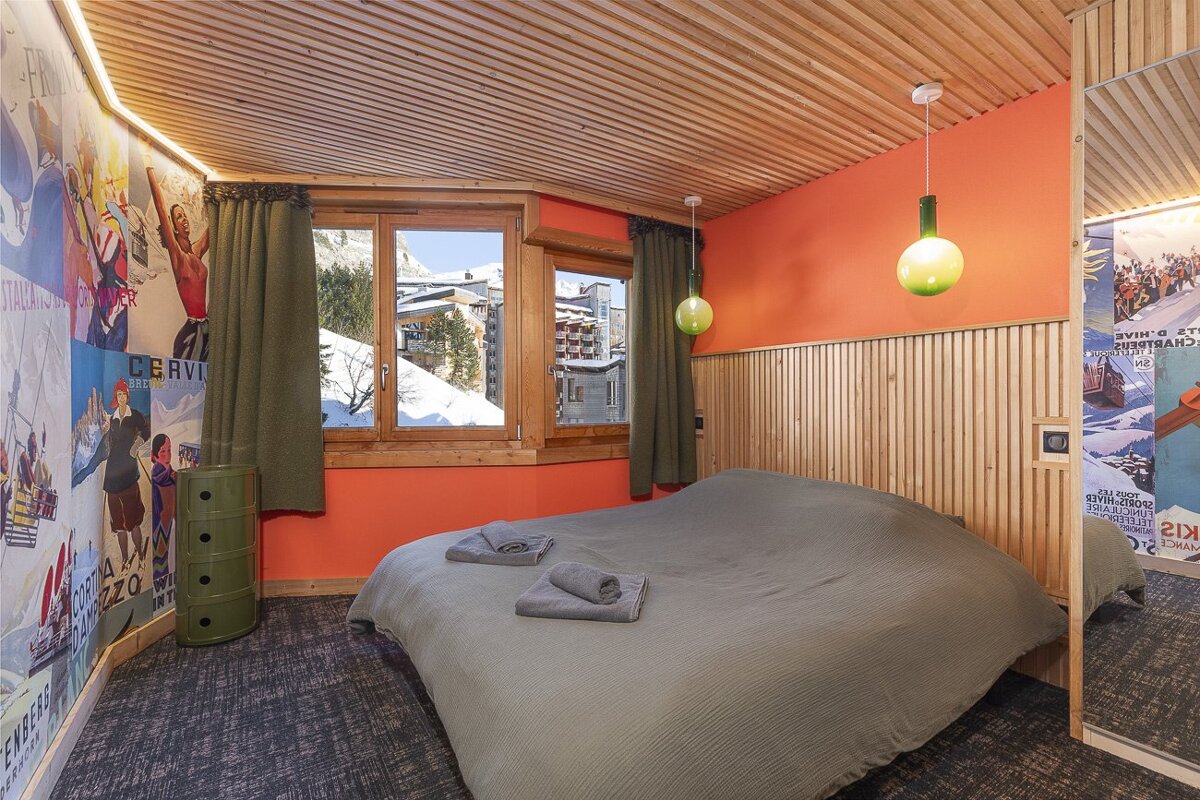 Cozy bedroom with wooden paneling, orange walls, and a large bed. Features ski-themed wallpaper and a window with a mountain view.