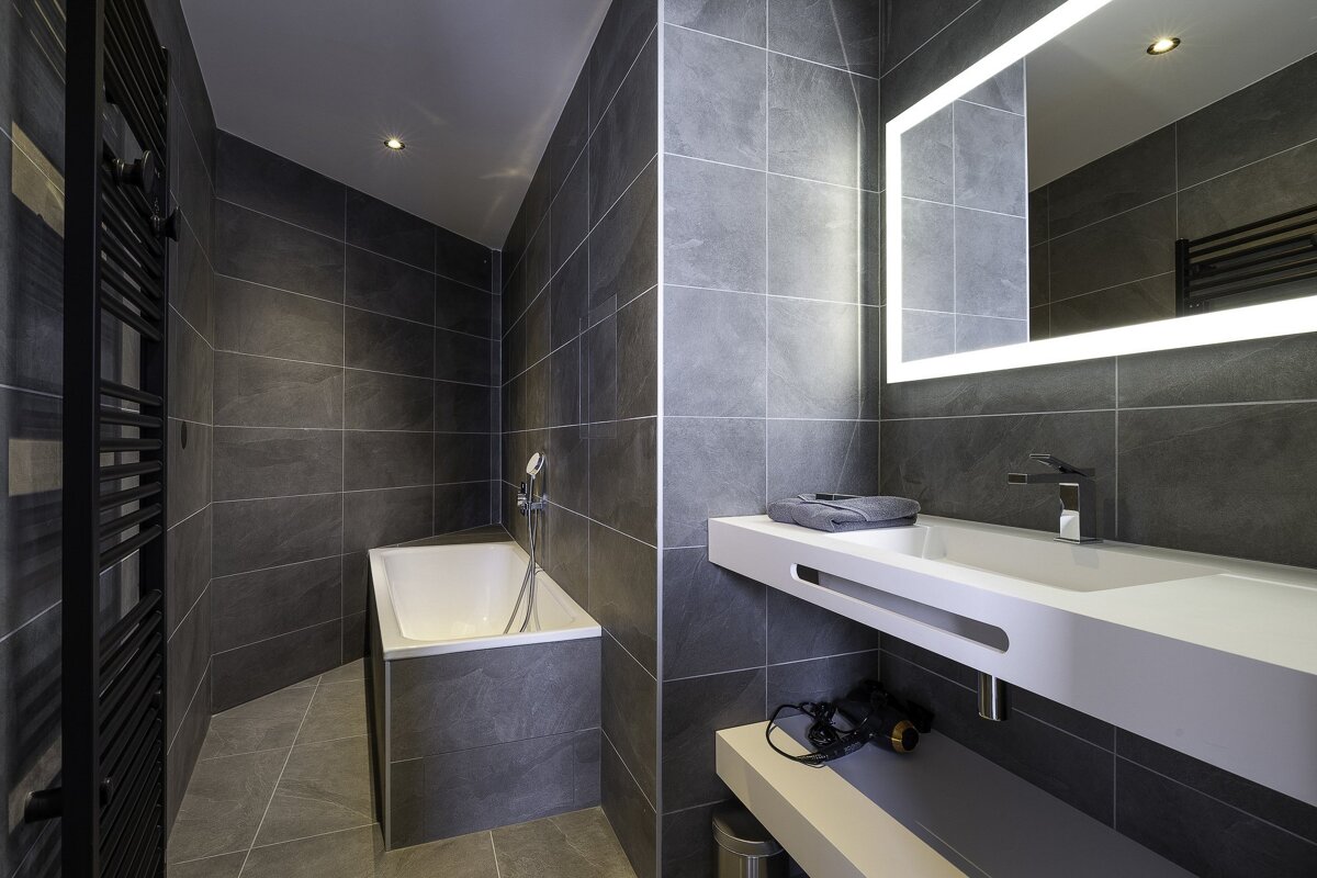 A modern, dark-tiled bathroom featuring a white sink under an illuminated mirror, and a white bathtub nestled in a corner.