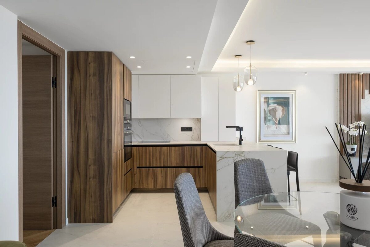A modern open-plan apartment with wood and white kitchen cabinets, a marble island, and a dining table with grey chairs.
