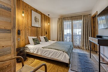 A cozy bedroom with a prominent wood-paneled wall, large bed, decorative pillows, and sheer curtains leading to a balcony.