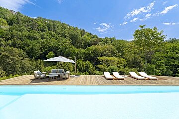 Modern infinity pool with a wooden deck, lounge chairs, and outdoor seating, set against a lush green hillside under a blue sky.