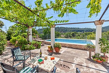 A table with a bottle of wine sits under a pergola overlooking a swimming pool