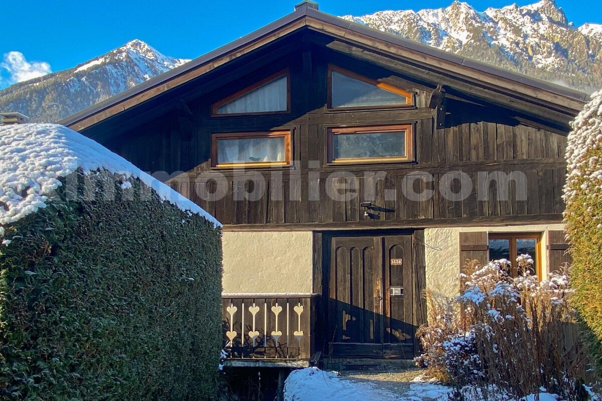 A dark wooden chalet against snowy mountains, bathed in sunlight. Snow covers hedges and ground, contrasting with the dark wood and white walls.