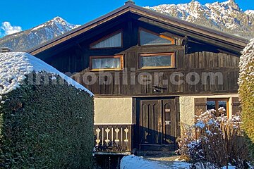 A dark wooden chalet against snowy mountains, bathed in sunlight. Snow covers hedges and ground, contrasting with the dark wood and white walls.