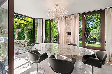Elegant dining room with an oval marble table, grey swivel chairs, and a chandelier, surrounded by windows showcasing a lush green garden.