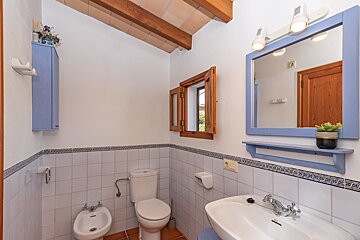 A rustic bathroom features white and blue tiled walls, exposed wooden beams, a toilet, bidet, sink, and a blue-framed mirror.