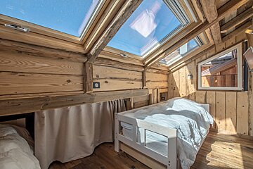 A cozy, wood-paneled bedroom with a large skylight showing a blue sky. There's a white bed on the right and another bed partially hidden on the left.