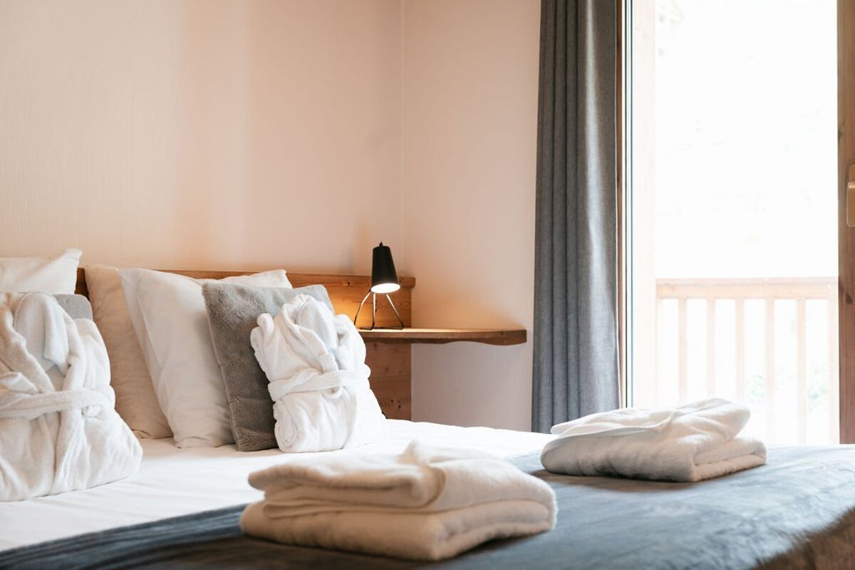 Cozy bedroom with white bed, bathrobes, and folded towels. A black lamp sits on a wooden nightstand beside a bright window with a balcony.