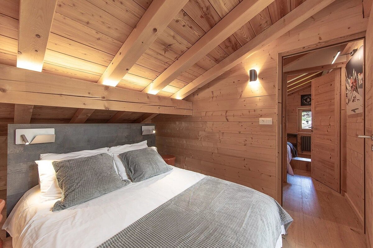 Cozy wood-paneled attic bedroom with slanted wooden ceiling, modern lighting, a large bed with grey bedding, and a doorway leading to another room.