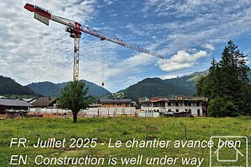 A construction site with a large crane, buildings under development, green fields, and mountains. Text: Construction is well under way.