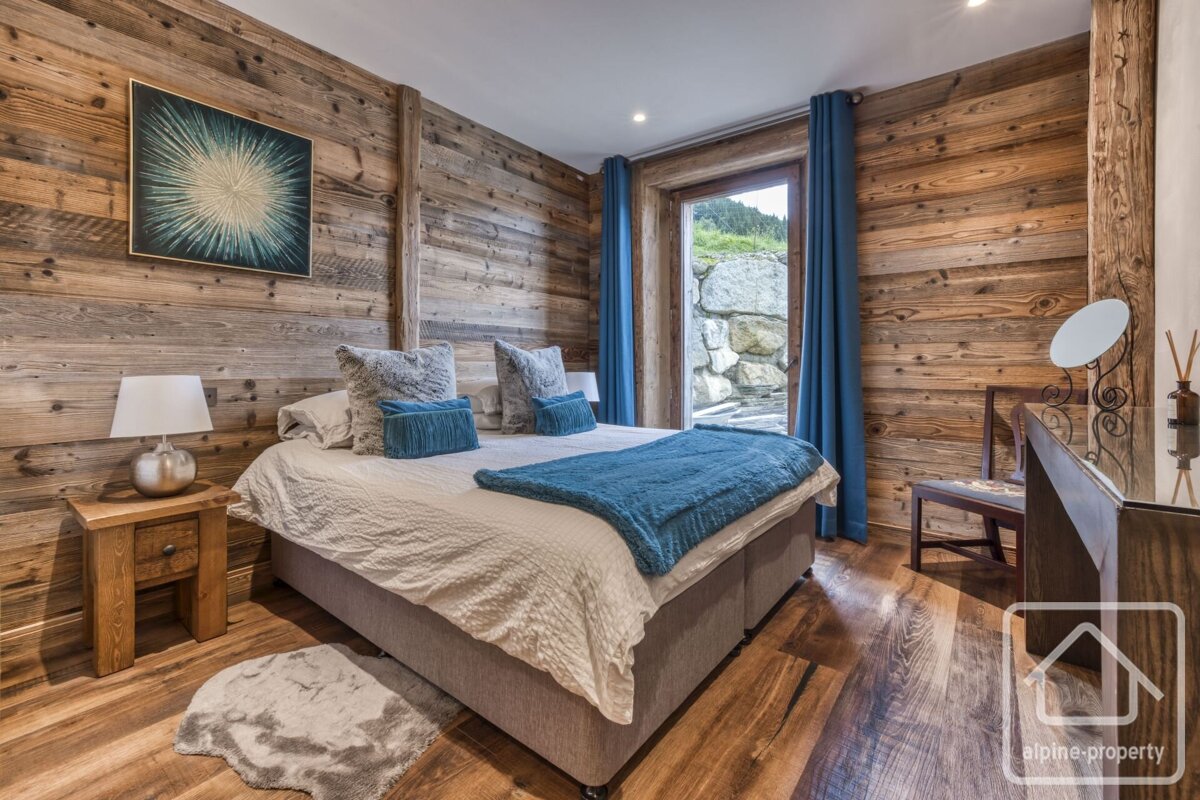 A cozy, rustic bedroom featuring warm wooden walls/floor, a large bed with blue linens, and a door opening to a stone outdoor area.