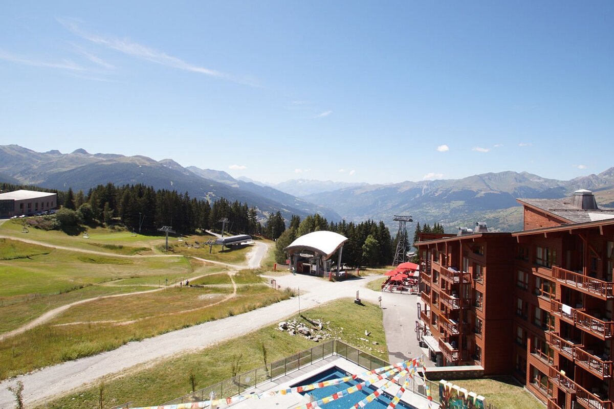 Sunny summer view of an Alpine resort, featuring a large brown building, ski lifts, green slopes, a pool, and distant mountains.
