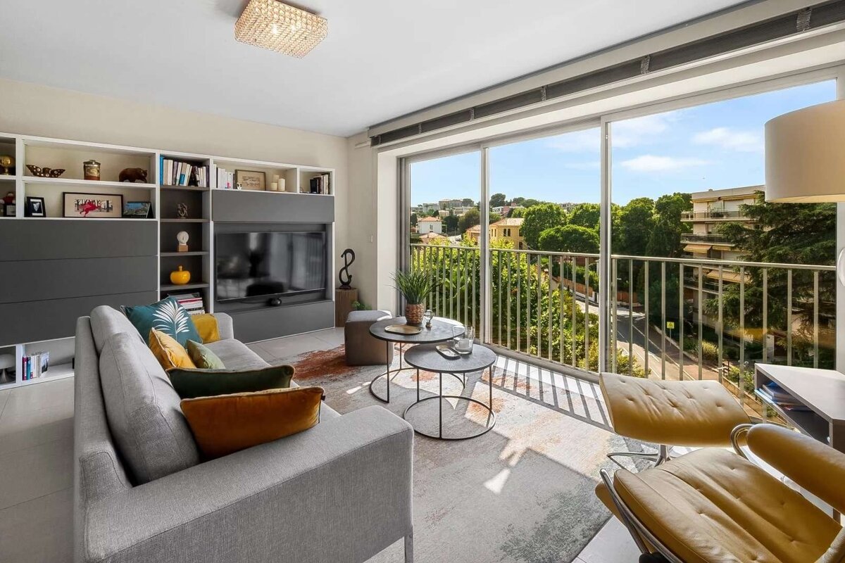 Bright, modern living room with a gray sofa, yellow armchair, built-in TV unit, and a balcony offering a verdant city view.