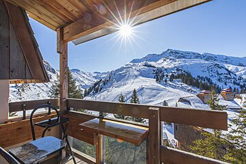 A bright sun shines over snowy mountains and ski slopes, seen from a rustic wooden balcony.