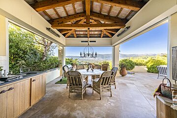 A beautiful covered outdoor patio with an al fresco dining set, outdoor kitchen, exposed wooden ceiling, and a stunning panoramic view of a scenic valley.