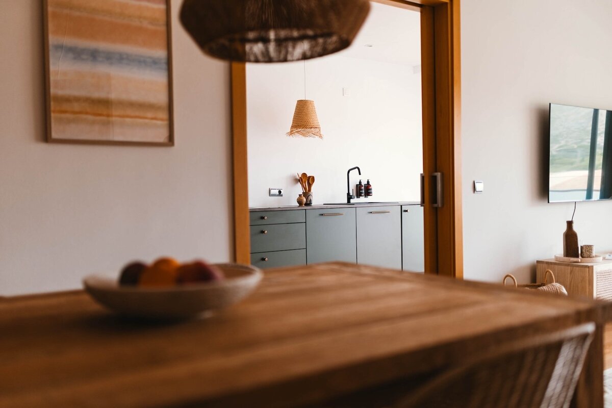 A cozy interior featuring a blurred wooden table, opening to a minimalist kitchen with sage green cabinets, black faucet, and a TV on the side.