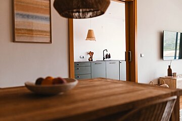 A cozy interior featuring a blurred wooden table, opening to a minimalist kitchen with sage green cabinets, black faucet, and a TV on the side.