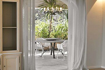 A view through sheer curtains to an outdoor patio with a dining table, modern white chairs, lush green plants, and a black chandelier hanging overhead.