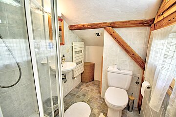 A rustic attic bathroom with light gray tiles, featuring a shower cabin, sink, toilet, and prominent wooden beams across the sloped ceiling.