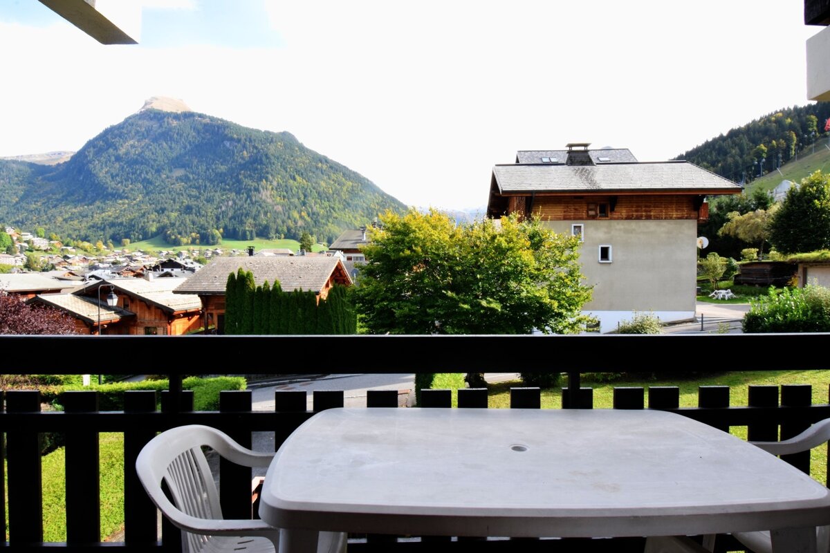 Balcony view over a charming mountain village and a forested peak. A white plastic table and chair are in the foreground.