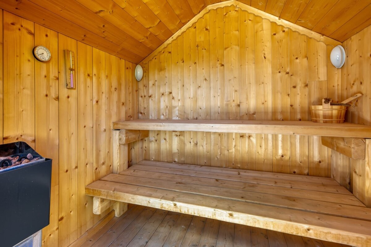 An empty, traditional wooden sauna with two benches, a heater, a thermometer, and a wooden bucket and ladle.