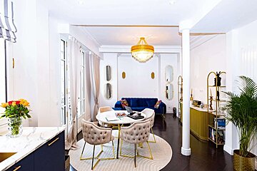 Stylish white and gold living/dining room with a blue velvet sofa, round table, tufted chairs, dark wood floors, and bright accents.